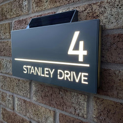 A solar-powered LED illuminated house number sign mounted on a brick wall, displaying the number 4 and the text 'STANLEY DRIVE'.